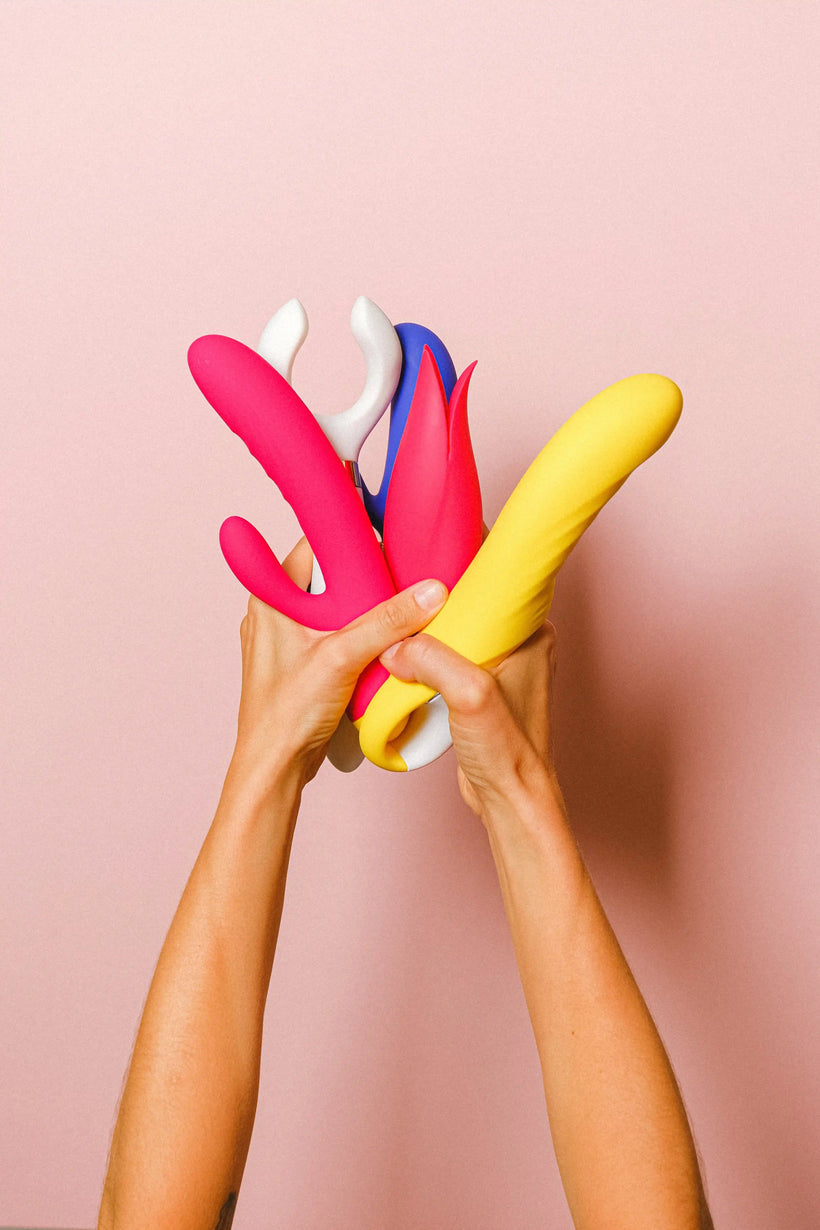 Dos manos sosteniendo varios vibradores de colores —rosa, amarillo y blanco— sobre fondo rosa pálido.