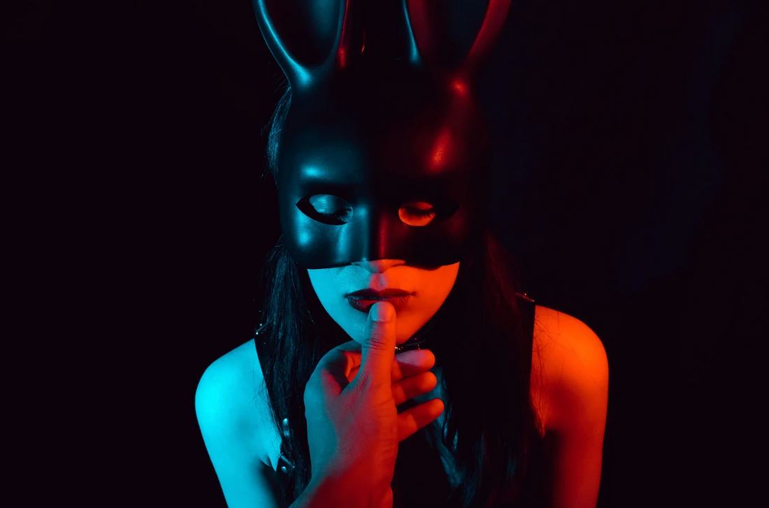 Personne portant un masque noir style lapin, éclairage bleu et rouge, doigt sur les lèvres (signe de silence).