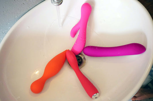 Trois sextoys colorés (rose, violet, orange) dans un lavabo près de la bonde, eau qui coule