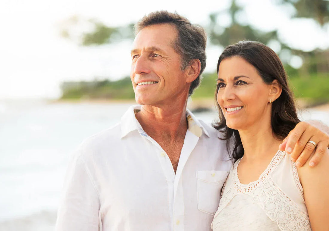 Couple mûr souriant au bord de la mer, bras autour des épaules, regard vers l’horizon.