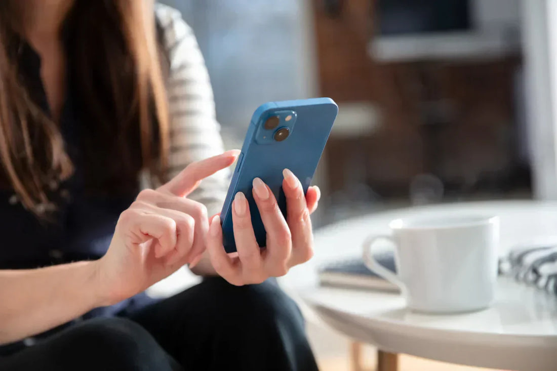 Gros plan d’une personne utilisant un smartphone bleu, assise près d’une table avec une tasse blanche.