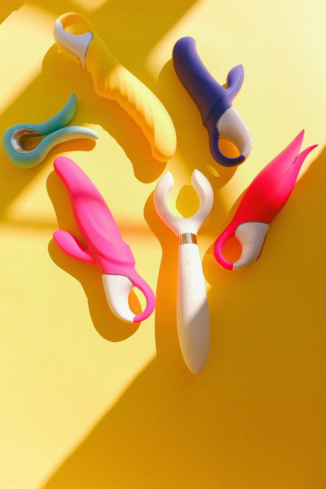 Six sextoys colorés disposés sur un fond jaune lumineux, avec de fortes ombres.