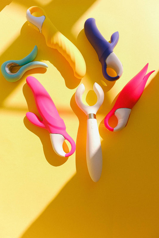 Six sextoys colorés disposés sur un fond jaune lumineux, avec de fortes ombres.