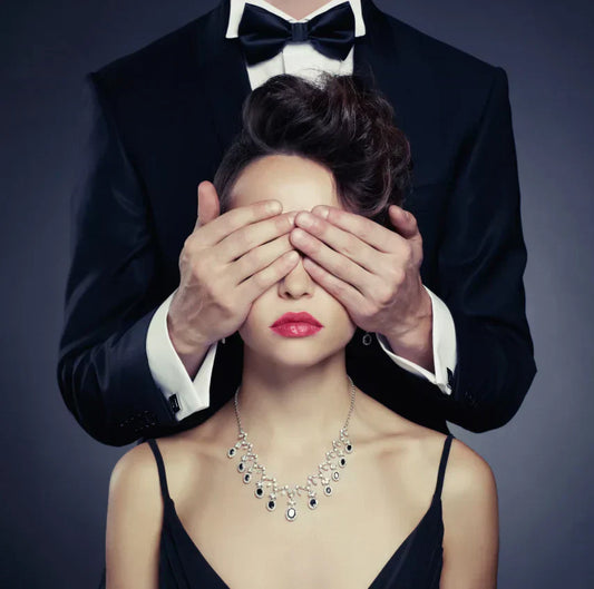 Gros plan sur une femme élégante en robe de soirée et collier de diamants, les yeux bandés par les mains d'un homme en smoking et nœud papillon.