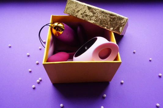 Boîte cadeau ouverte sur fond violet contenant plusieurs sextoys, avec couvercle doré posé et petites billes décoratives.