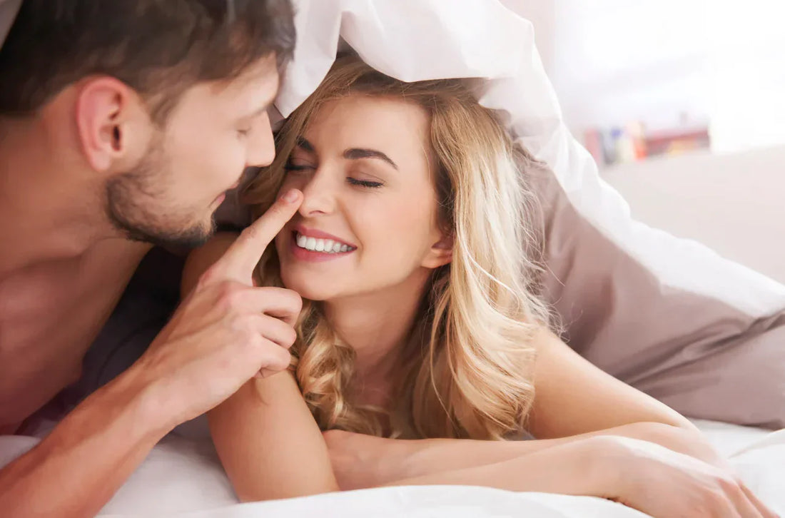 Couple allongé sous une couette, geste tendre et sourires.