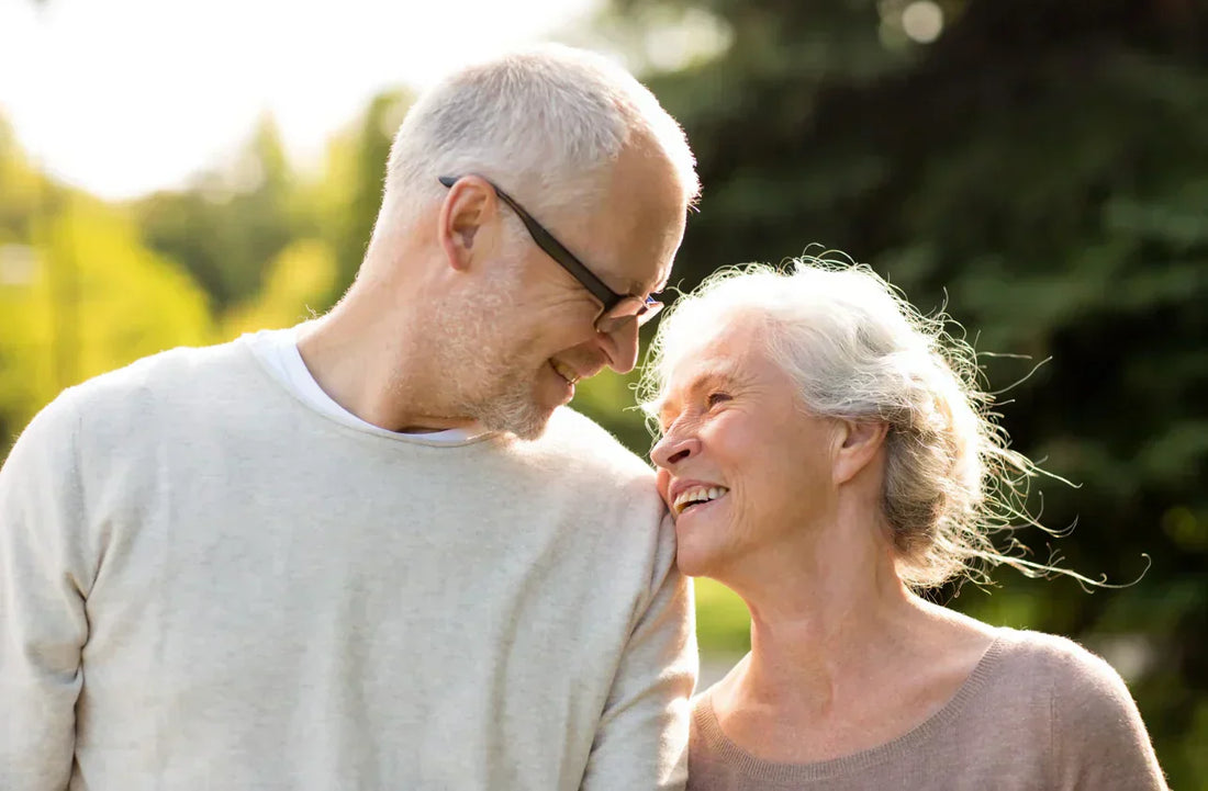 Couple senior souriant, se regardant tendrement lors d’une promenade en extérieur, lumière douce en arrière-plan.