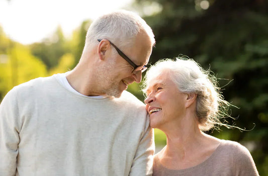 Couple senior souriant, se regardant tendrement lors d’une promenade en extérieur, lumière douce en arrière-plan.