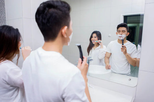 Couple dans une salle de bain devant le miroir : elle se brosse les dents, lui se rase avec de la mousse.