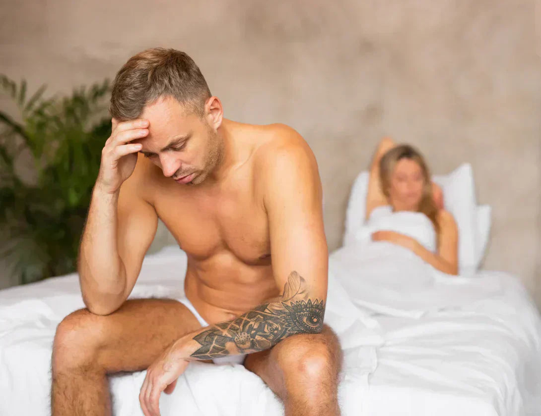 Homme assis au bord du lit, tête dans la main, partenaire floue allongée derrière.