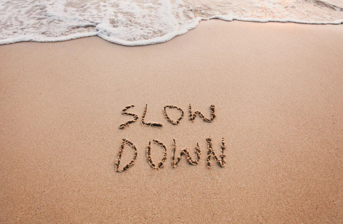 Mots « SLOW DOWN » écrits dans le sable sur une plage, avec les vagues en haut de l’image.