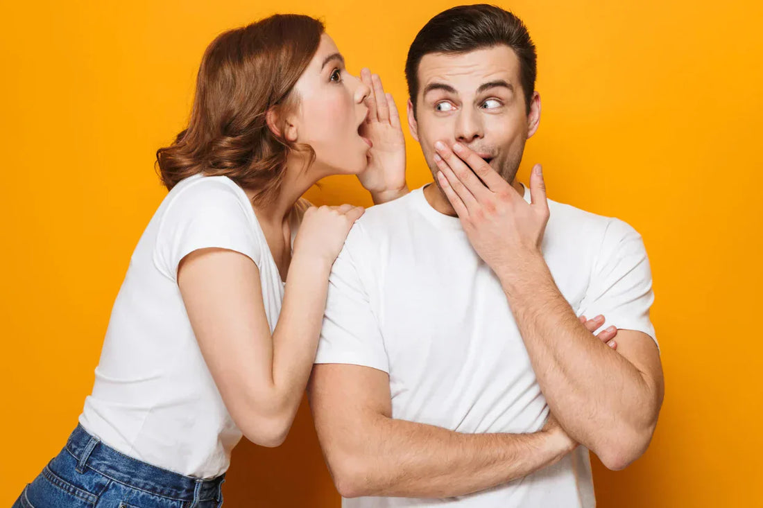 Femme chuchotant à l’oreille d’un homme surpris, sur fond orange.
